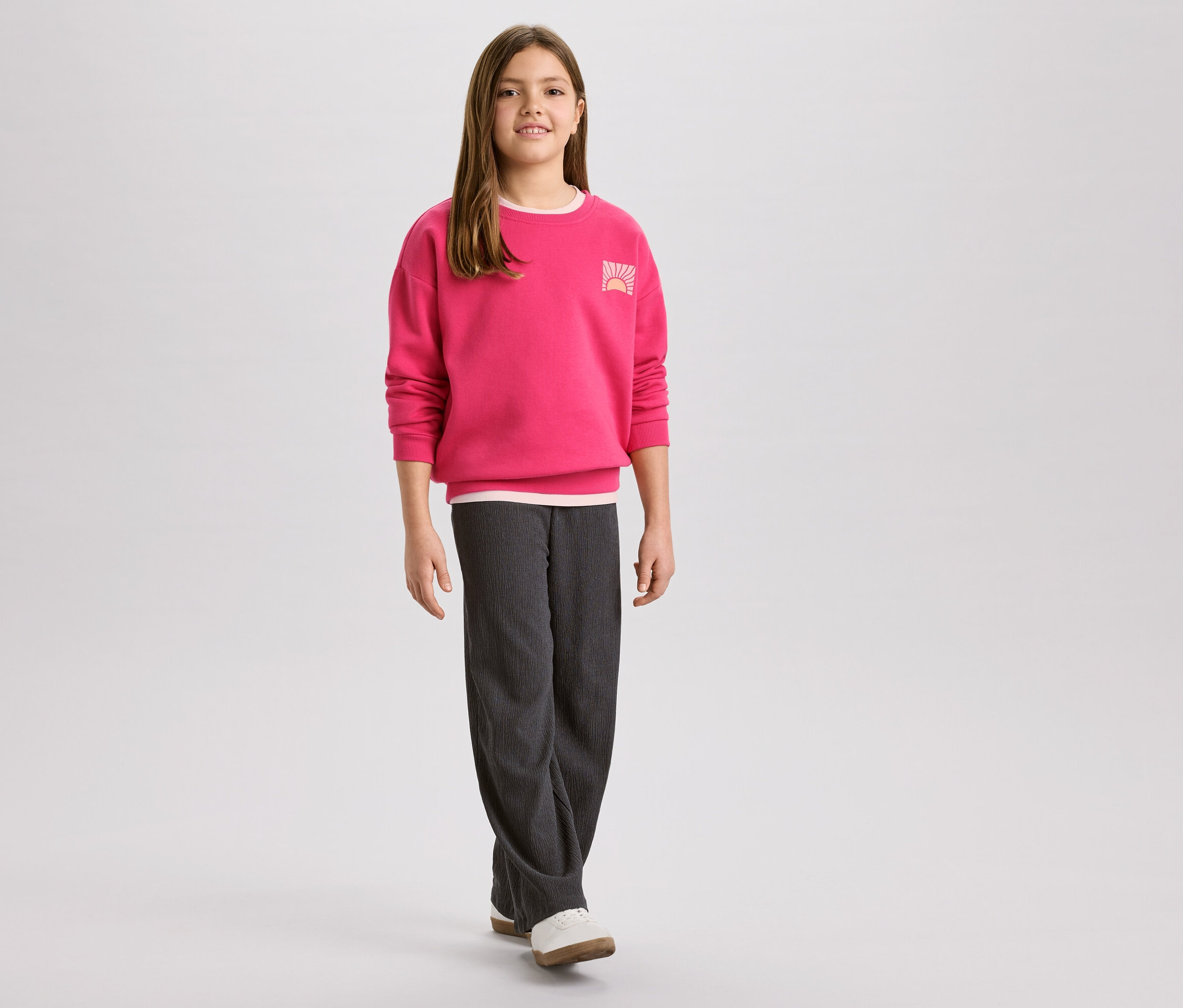 Lány rózsaszín pulóverben, napkelte logóval és szürke kordbársony nadrágban, hozzá fehér cipő.