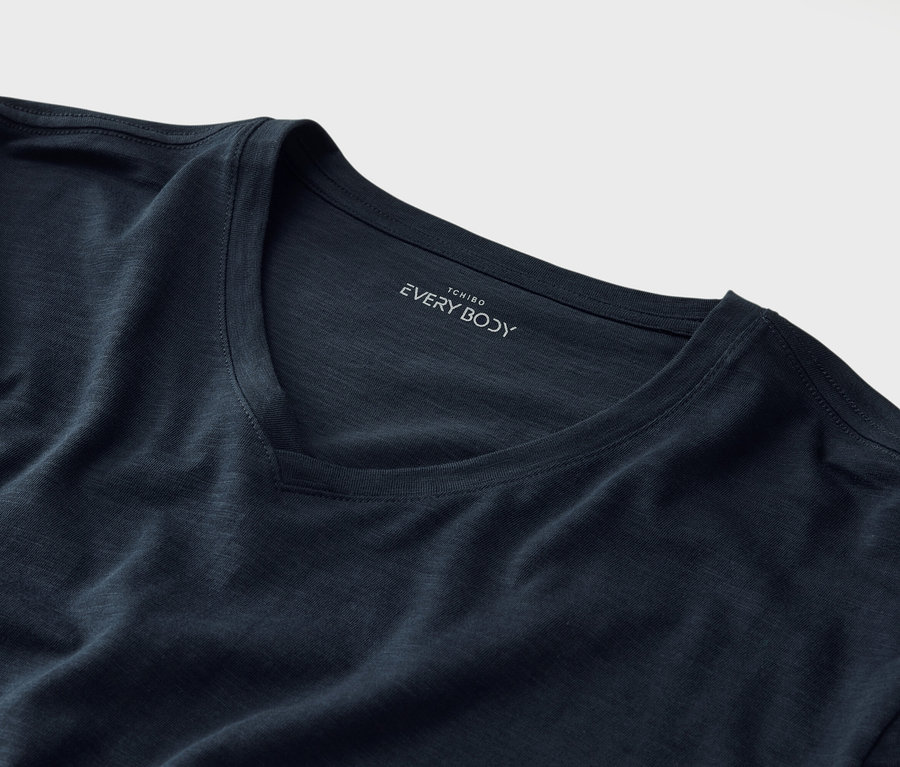 Egy sötétkék, V-nyakú póló közeli képe, a gallér alatt a „Tchibo EVERY BODY” felirattal.