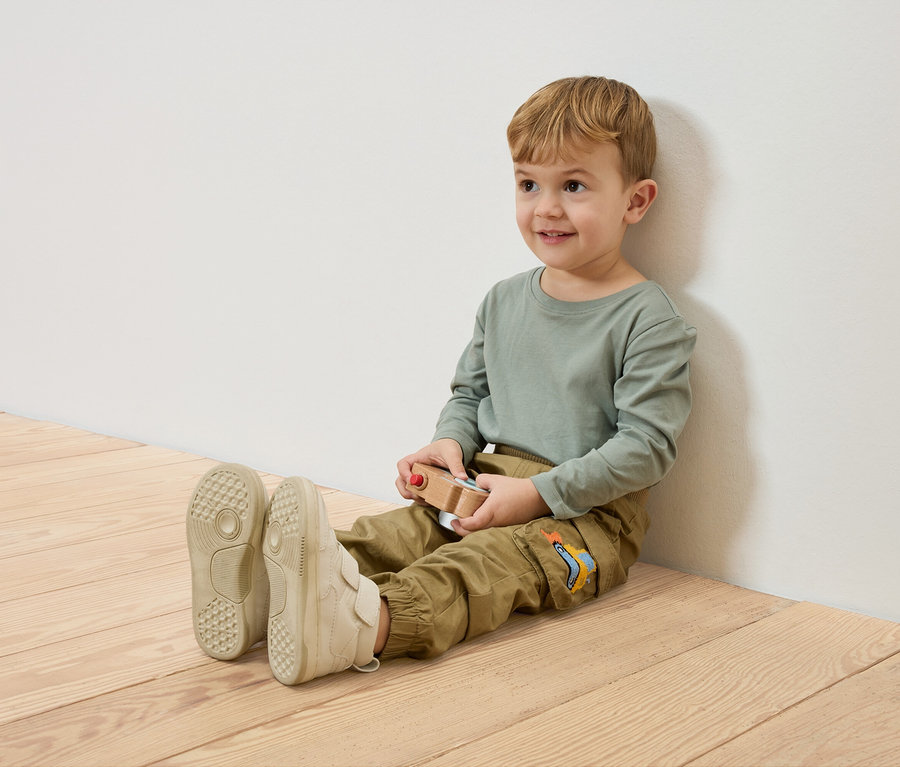 Egy mosolygó fiú ül a fapadlón, egy fehér falnak támaszkodva, egy fa játékautót tartva.