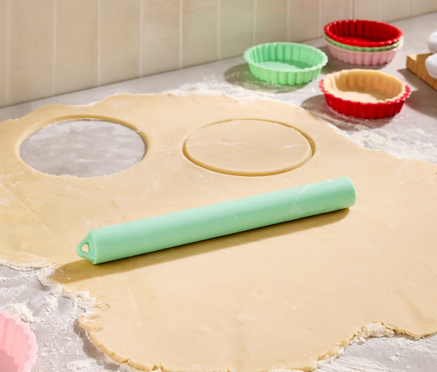 Lisztel szórt felületen kinyújtott tészta két kivágott körrel, egy zöld tésztanyújtó és színes piteformák láthatók.