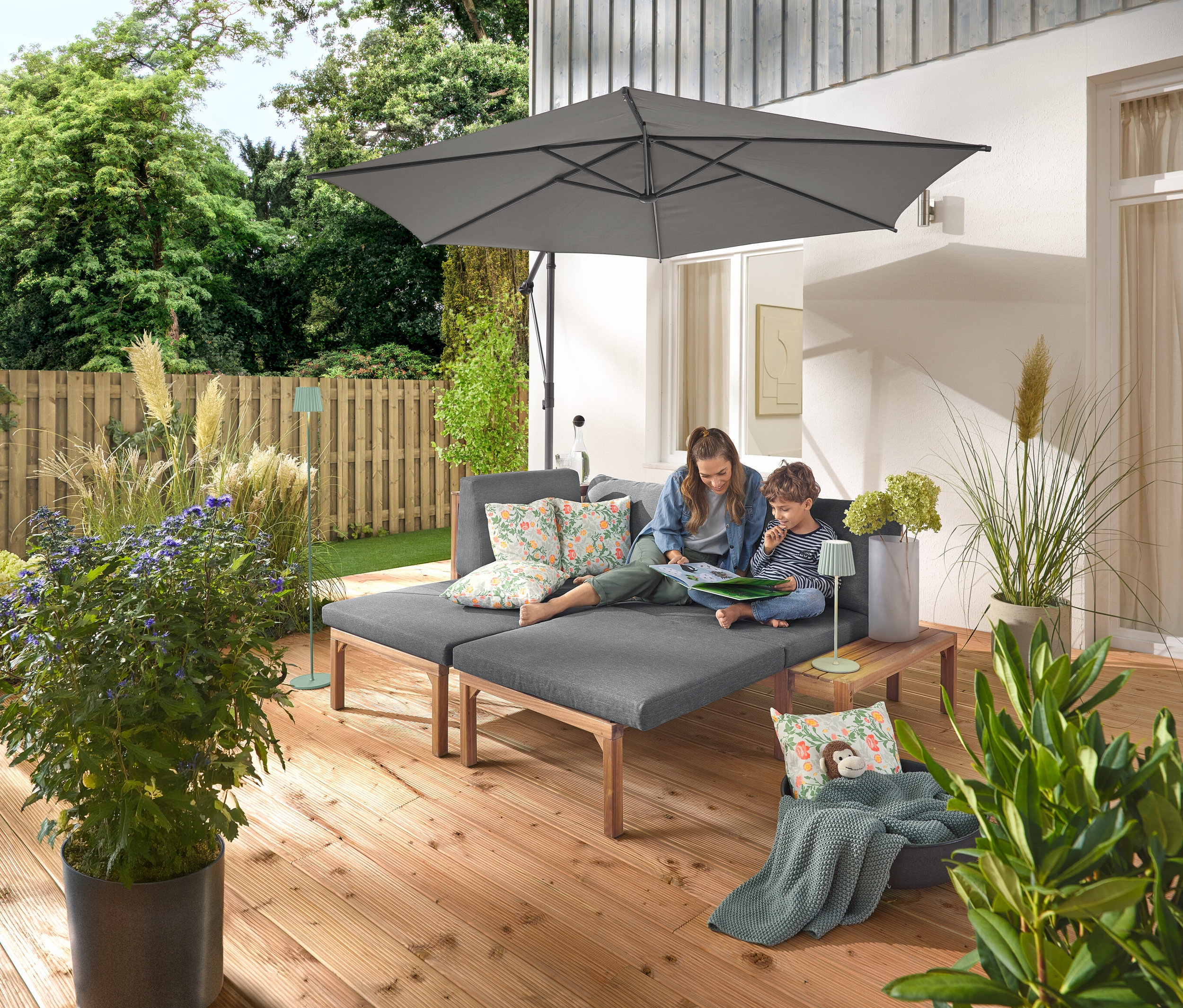 Egy nő és egy gyermek ül egy szürke kerti kanapén, külön kihúzható lábtartókkal. A kanapén két virágmintás kültéri párna található. Az antracit Leco napernyő állvánnyal és huzattal árnyékot biztosít. A kanapé melletti fa asztalon zsálya színű LED-es akkus kültéri asztali lámpa áll, a háttérben pedig egy zsálya színű LED-es akkus kültéri állólámpa látható.