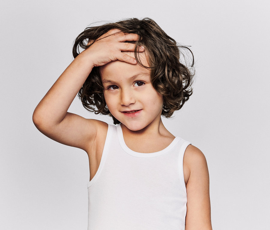 Portré egy göndör, barna hajú, fehér trikós gyermekről, aki a homlokára teszi a kezét.