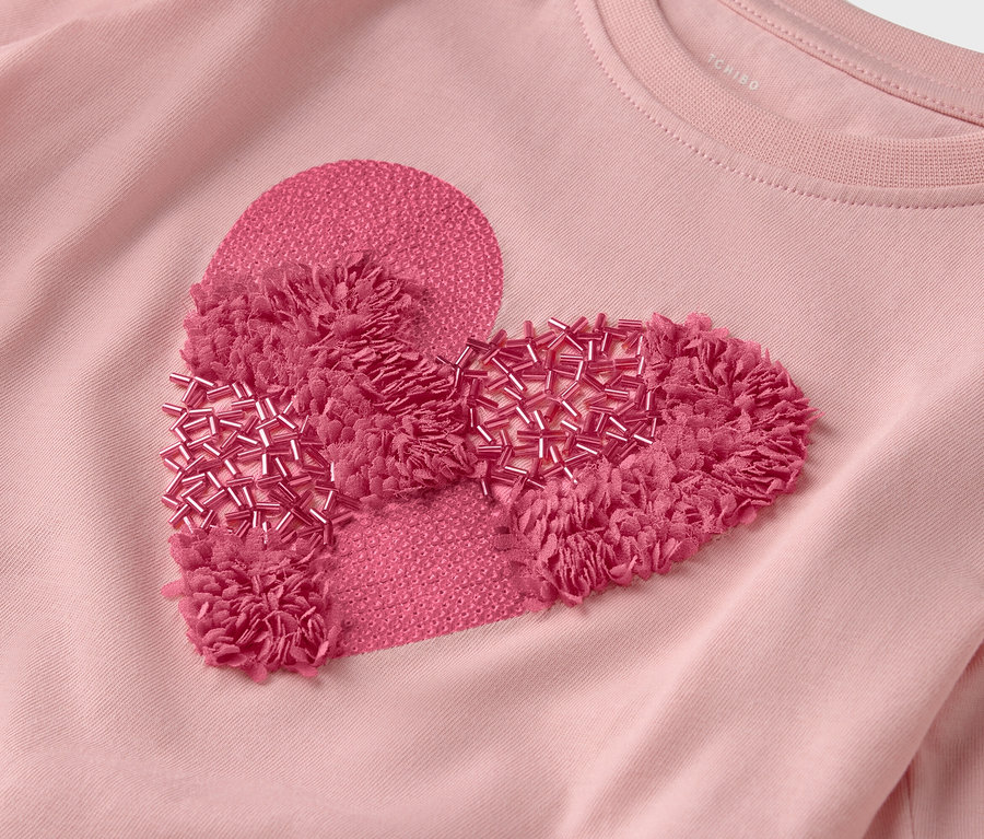 Egy rózsaszín póló közeli képe, szív alakú, rojtokkal és gyöngyökkel díszített folttal.