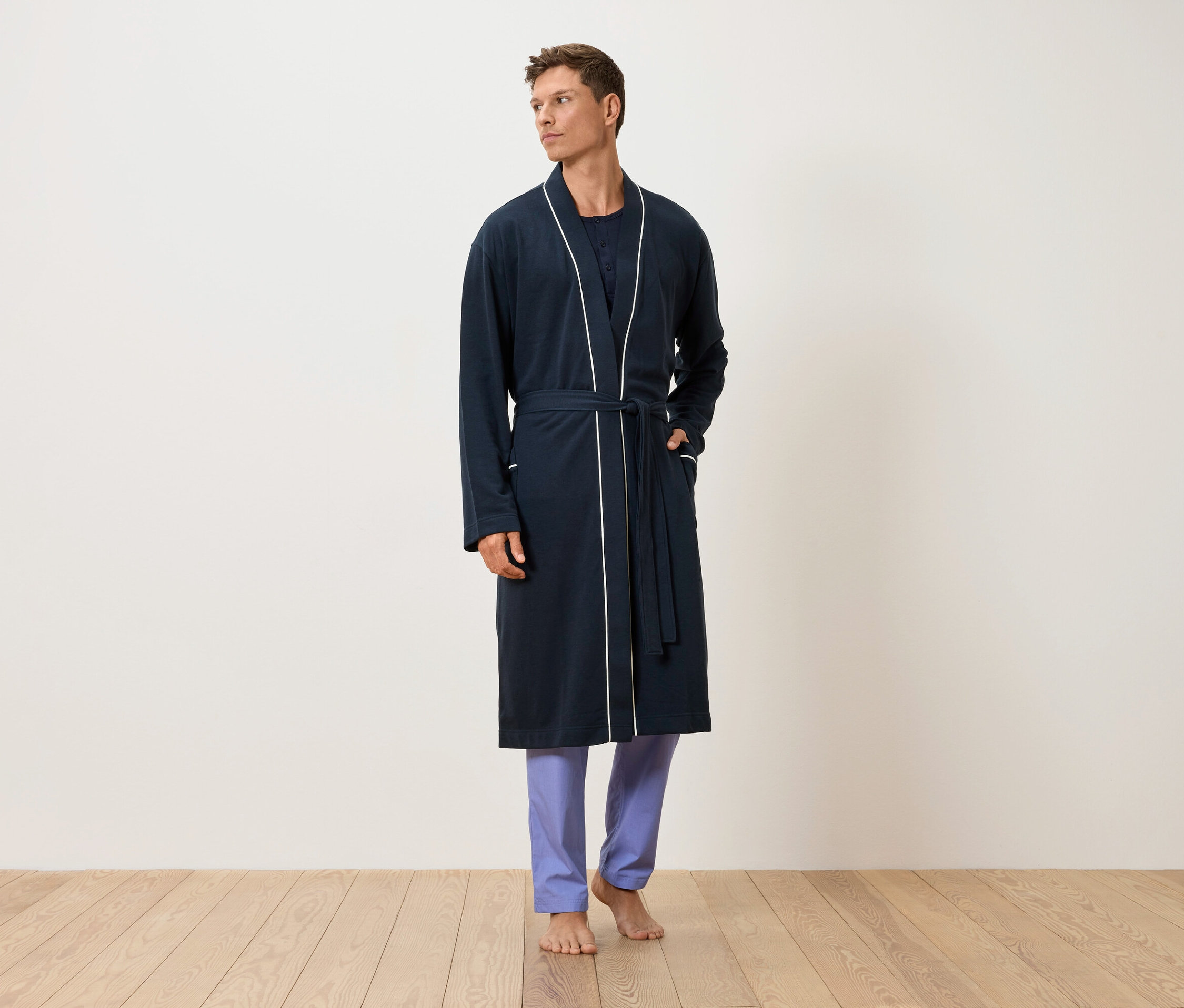 Egy férfi mezítláb áll egy fapadlón, sötétkék, fehér szegélyű köntösben és kék nadrágban.