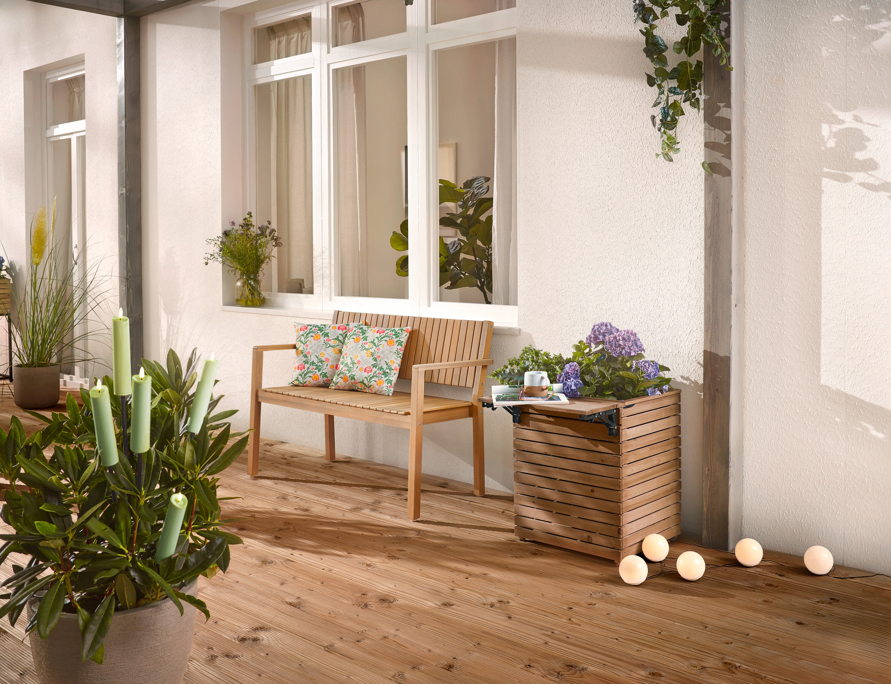 A fa teraszon egy fából készült pad párnákkal, egy virágláda virágokkal és egy cserepes növény gyertyákkal található.