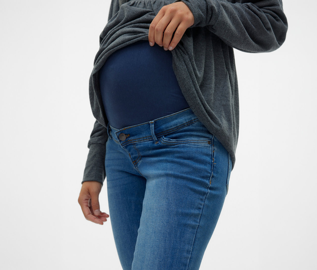 Nő húzza fel a szürke felsőt a hasa fölé, MAMALICIOUS kismama farmernadrágot és sötétkék kismama felsőt visel.