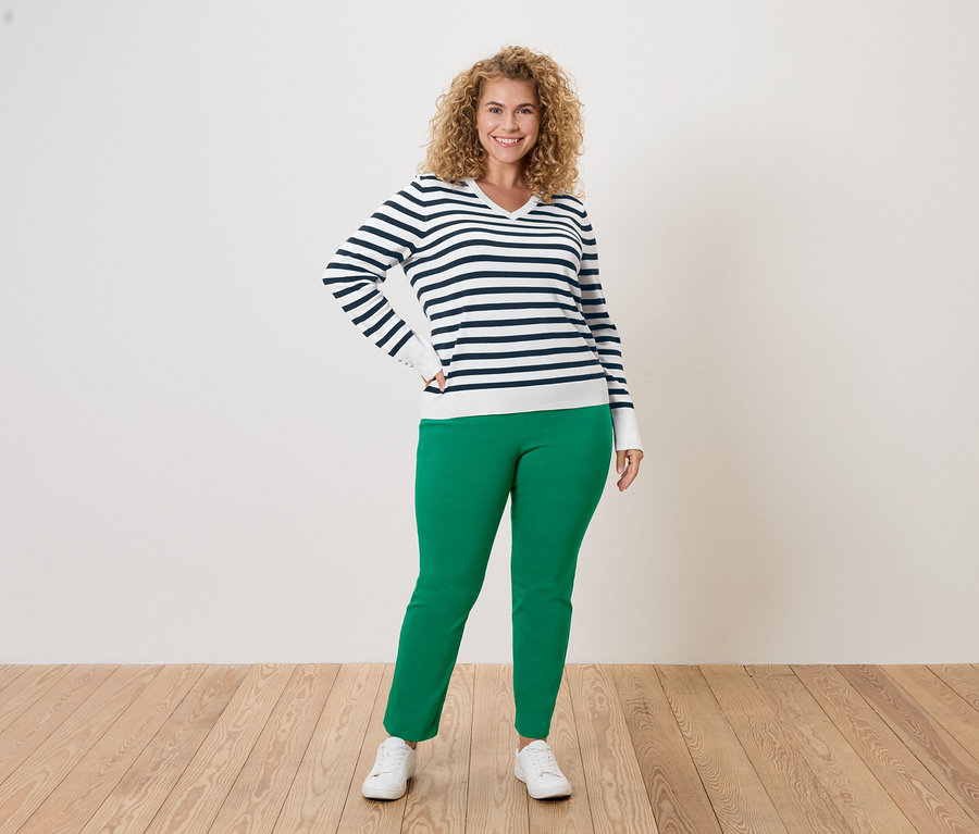 Göndör hajú nő kék-fehér csíkos pulóverben, zöld nadrágban és fehér tornacipőben pózol fehér háttér előtt.