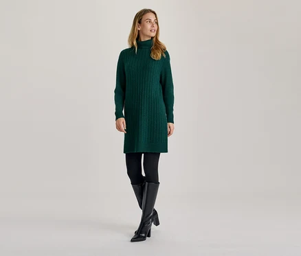 Nő zöld garbós kötött ruhában, fekete leggingsben és fekete térdig érő csizmában, oldalra nézve.