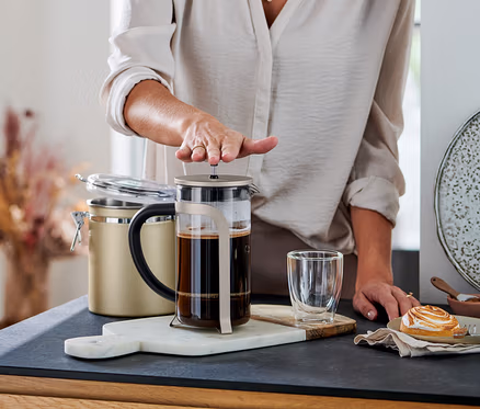 Egy személy lenyomja a kávéval teli pezsgőszínű dugattyús kávéfőző (800 ml, 6 csésze) dugattyúját. Mellette egy pohár és egy sütemény látható.