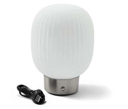 Egy LED-es akkumulátoros asztali lámpa üveg lámpabúrával egy fekete töltőkábel mellett áll.