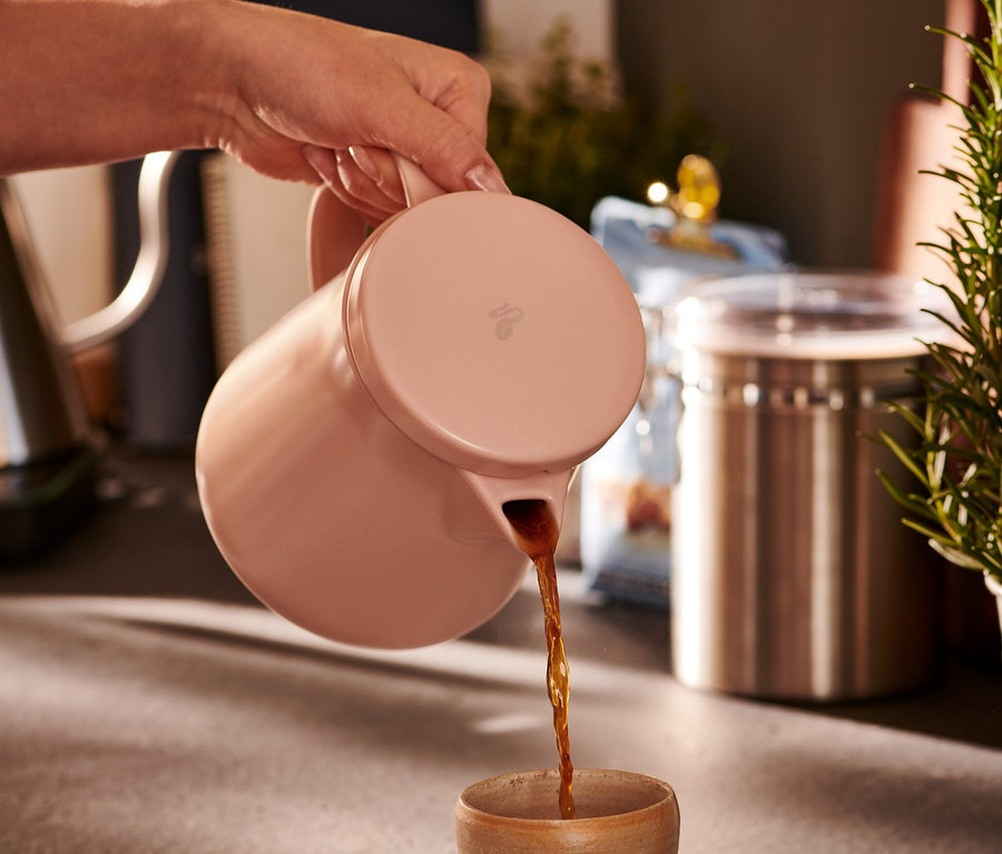 Egy kéz kávét önt egy rózsaszín kerámia kávéskannából egy csészébe.