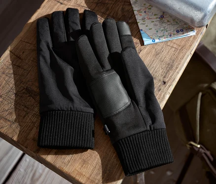Két pár fekete softshell kesztyű fekszik egymás mellett egy faasztalon. Mögöttük egy térkép és egy ezüstszínű kulacs látható.