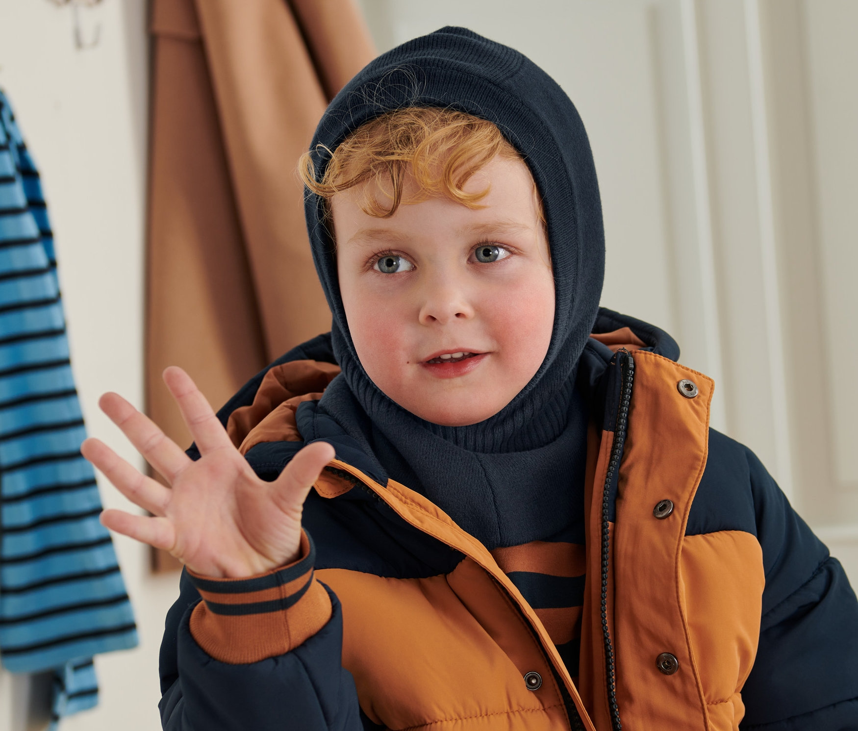 Fiú integet, sötétkék kisgyerek csősapkát és téli kabátot visel.