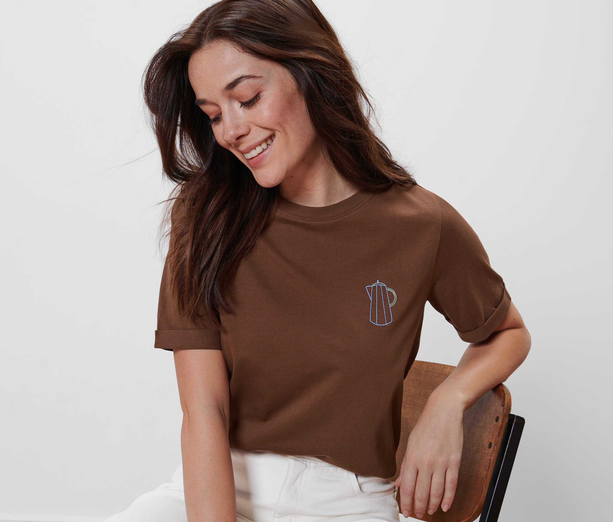 Nő ül egy széken, és hímzett, barna pólót visel.