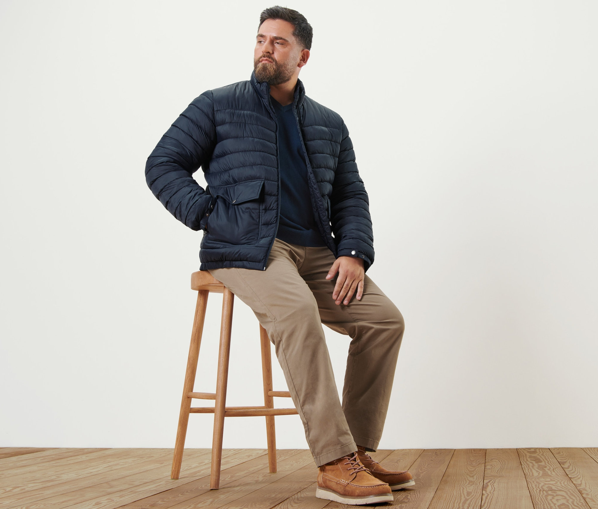 Férfi ül egy széken, barna chino nadrágot, tengerészkék V-nyakú pulóvert és sötétkék steppelt dzsekit visel.