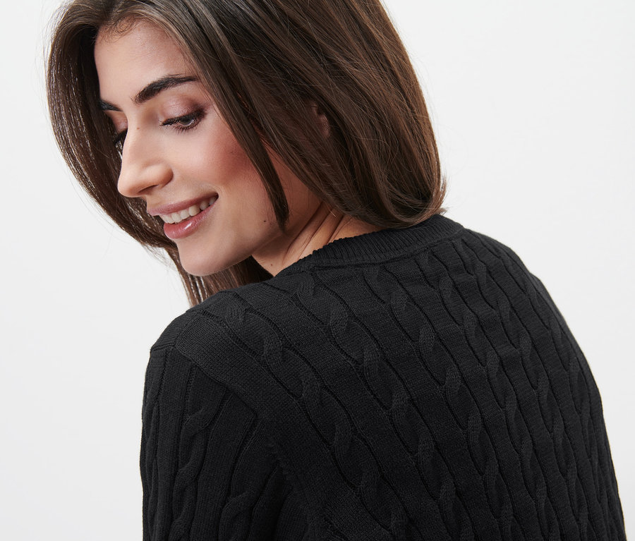 Mosolygó nő néz a válla fölött, fekete, fonott mintás kötött pulóvert visel.