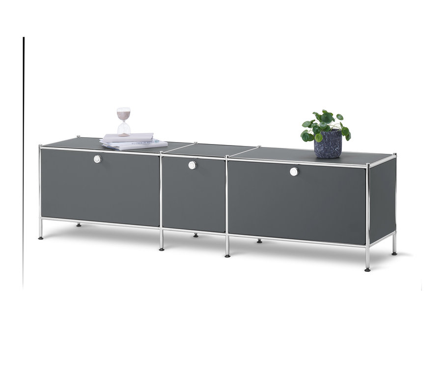 Egy szürke »CN3« alacsony komód 3 ajtóval fehér háttér előtt. A komódon cserepes növény, könyvek és homokóra található.