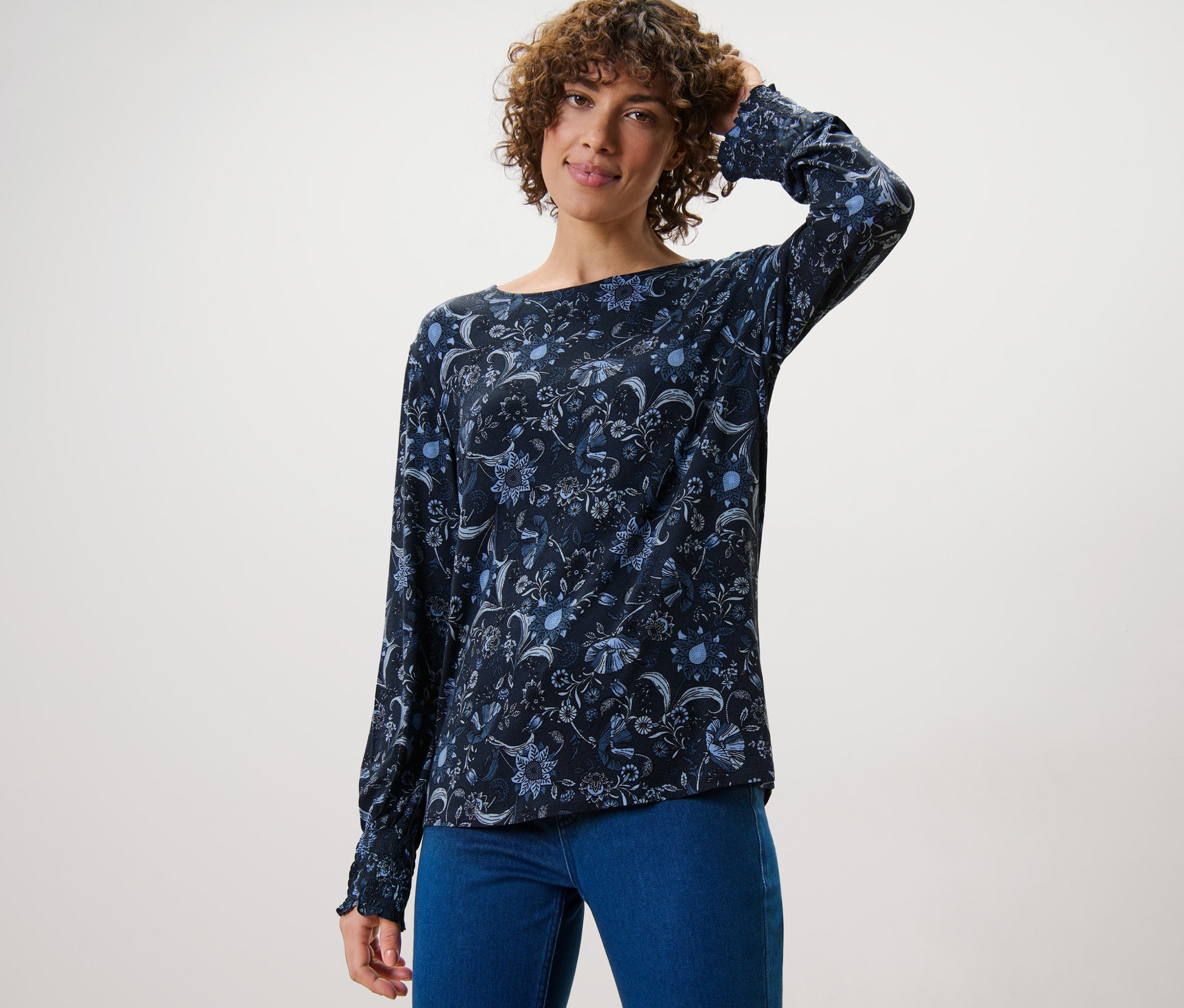 Nő pózol virágos hosszú ujjú pólóblúzban és farmerban.