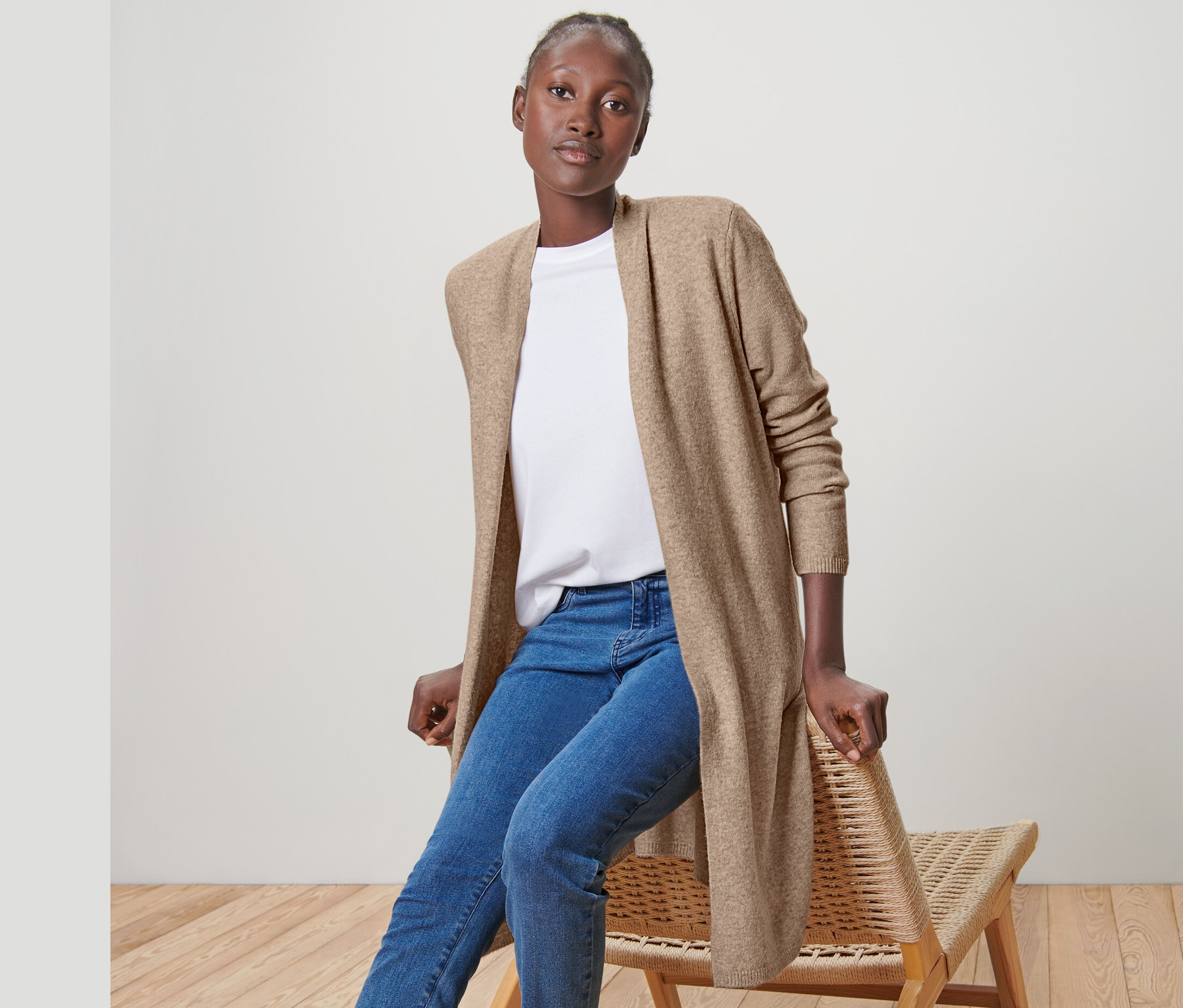 Nő ül egy széken, és egy bézs színű, gyapjúval készült női hosszú kardigánt, farmert és tornacipőt visel.