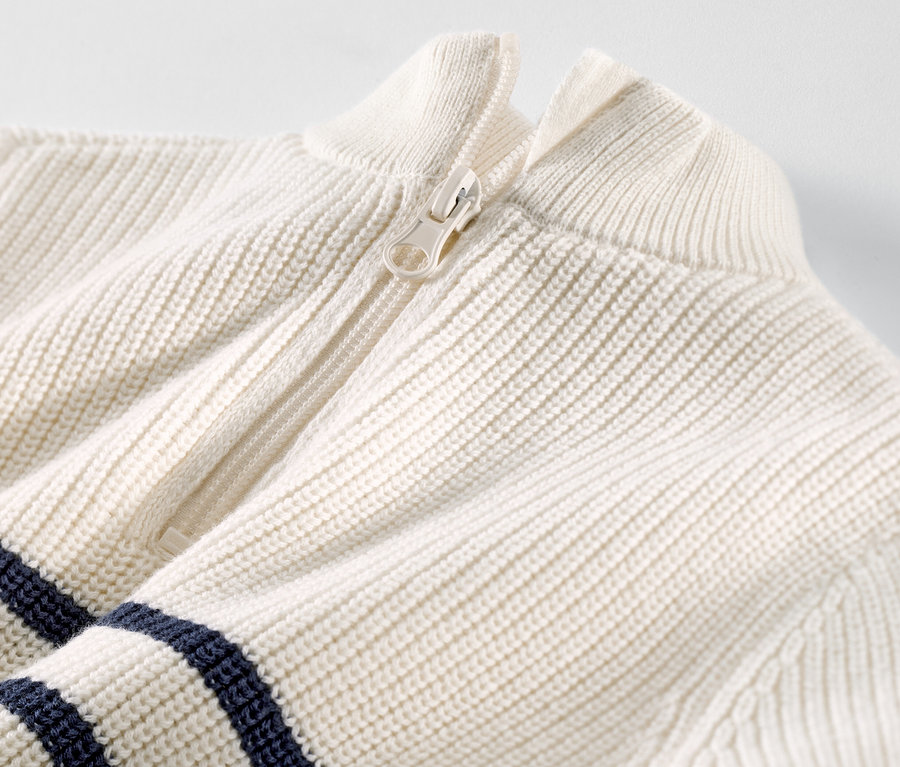 Közelkép egy fehér cipzáras, sötétkék csíkos állógalléros gyerekpulóverről.