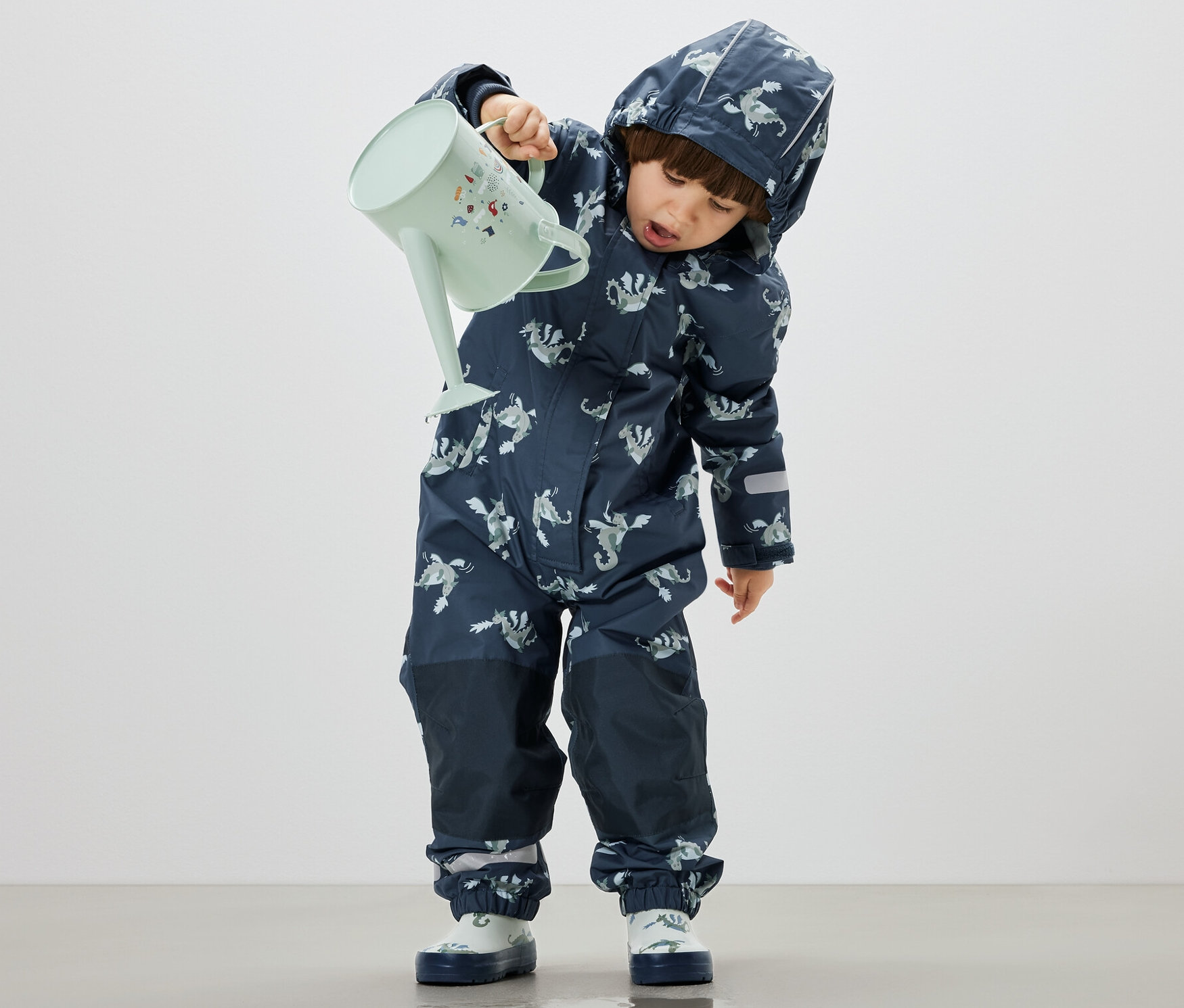 Fiú önti a vizet egy pasztellzöld kannából a padlóra, D eső elleni kezeslábast visel, lovagos.