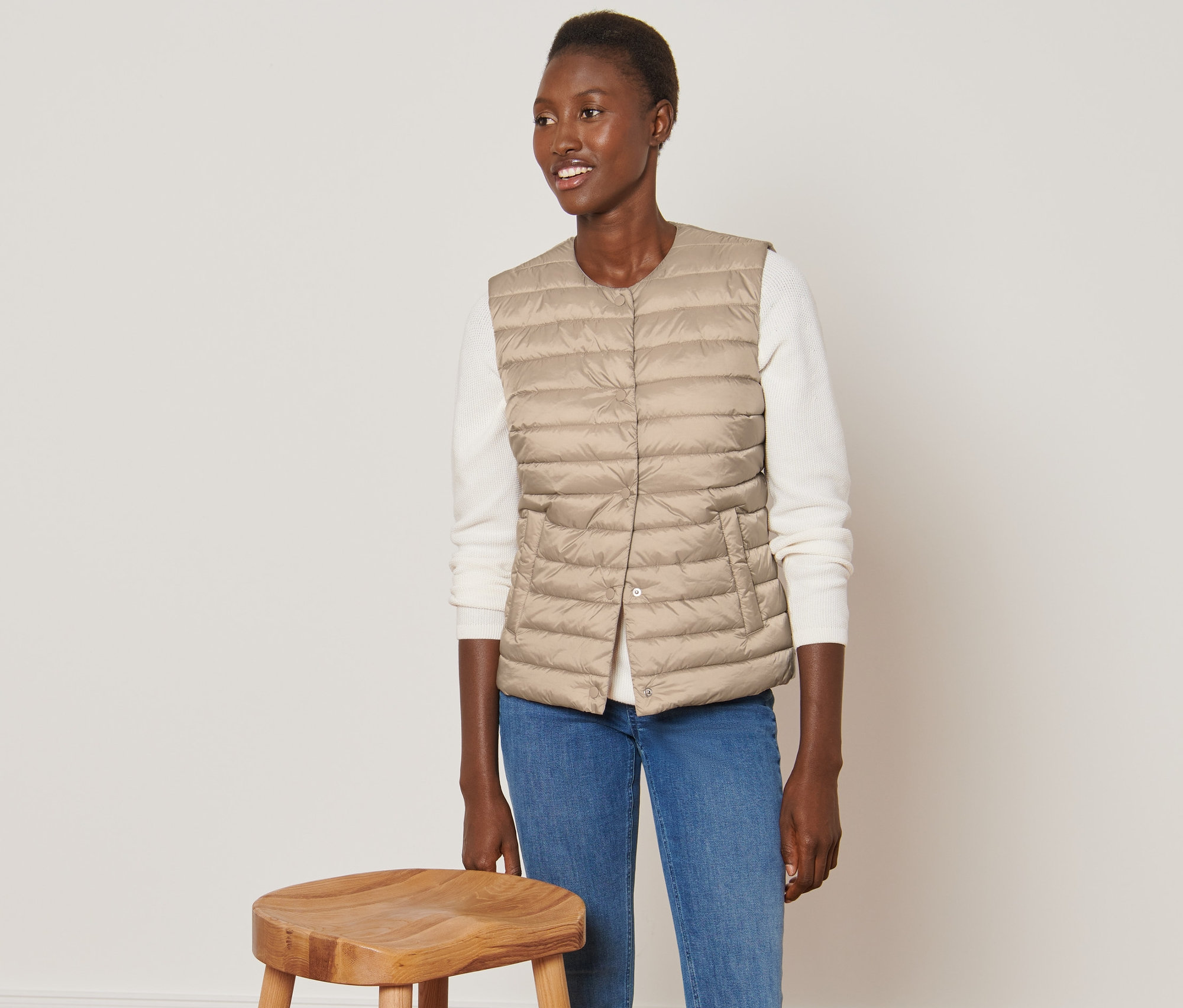 Nő visel bézs színű steppelt mellényt, és egy fa szék mellett áll.