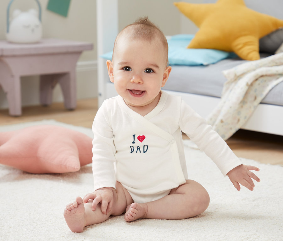 Mosolygó baba ül egy fehér szőnyegen, és egy fehér, "I love Dad" feliratú, hosszú ujjú body szettben lévő bodyt visel.