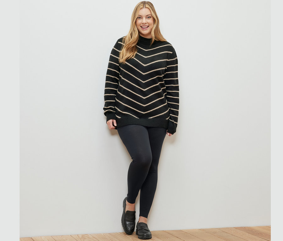 Nő pózol fekete csíkos kötött pulóverben, állógallérral és fekete leggingsben.