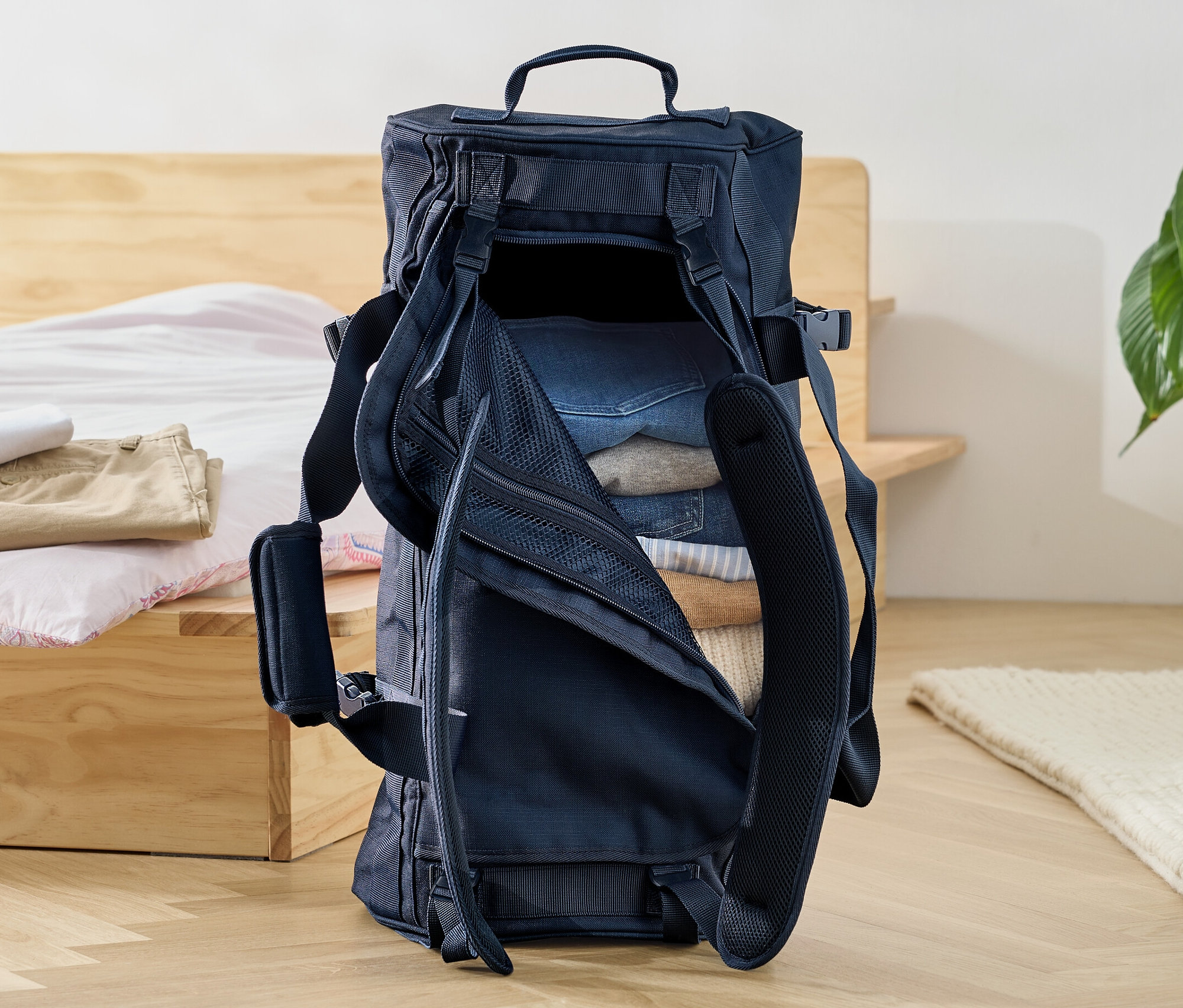 Egy nyitott utazótáska és hátizsák tele ruhákkal a padlón egy ágy mellett.