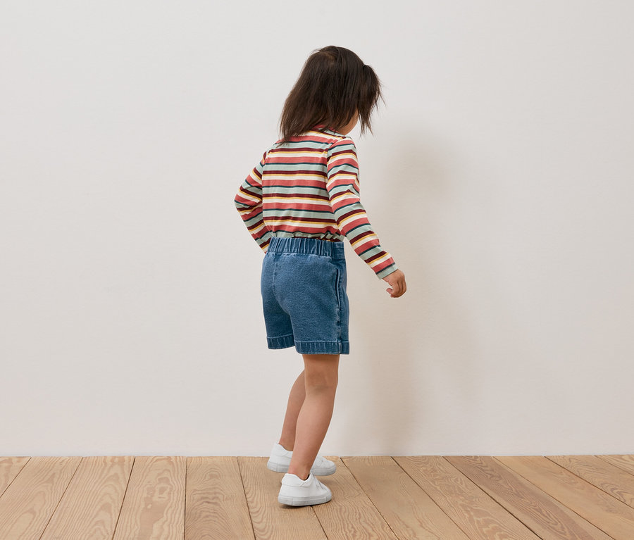Csíkos hosszú ujjú pólót és farmer rövidnadrágot viselő lány háttal áll a kamerának.