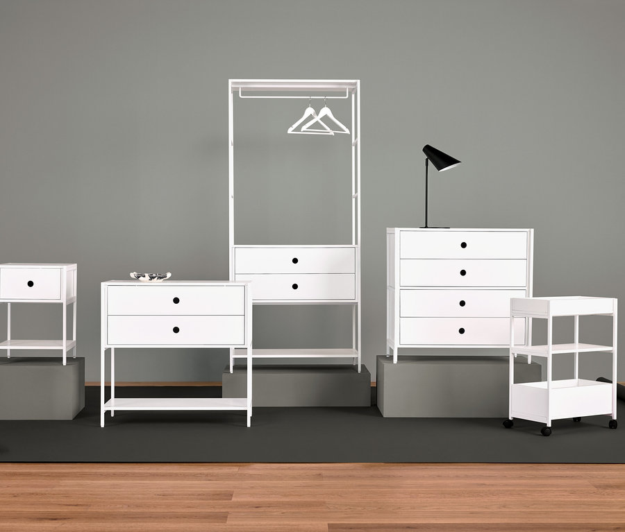 Fehér bútorok elrendezése éjjeliszekrénnyel, fiókos szekrénnyel, ruhaakasztóval, lámpával és polcokkal.