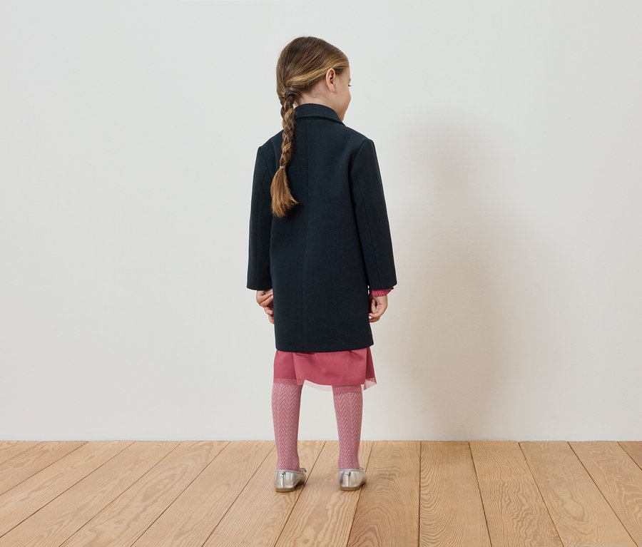 Egy kislány hátulról látható, sötétkék kabátot, rózsaszín ruhát, harisnyát és ezüst cipőt visel.