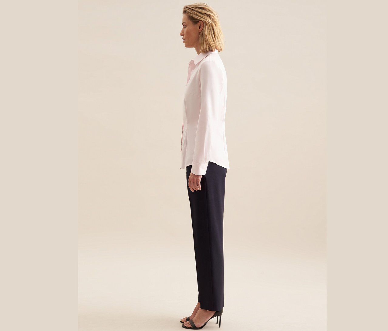 Egy nő oldalra állva rózsaszínű Seidensticker twill blúzt, sötétkék nadrágot és fekete, magas sarkú szandált visel.