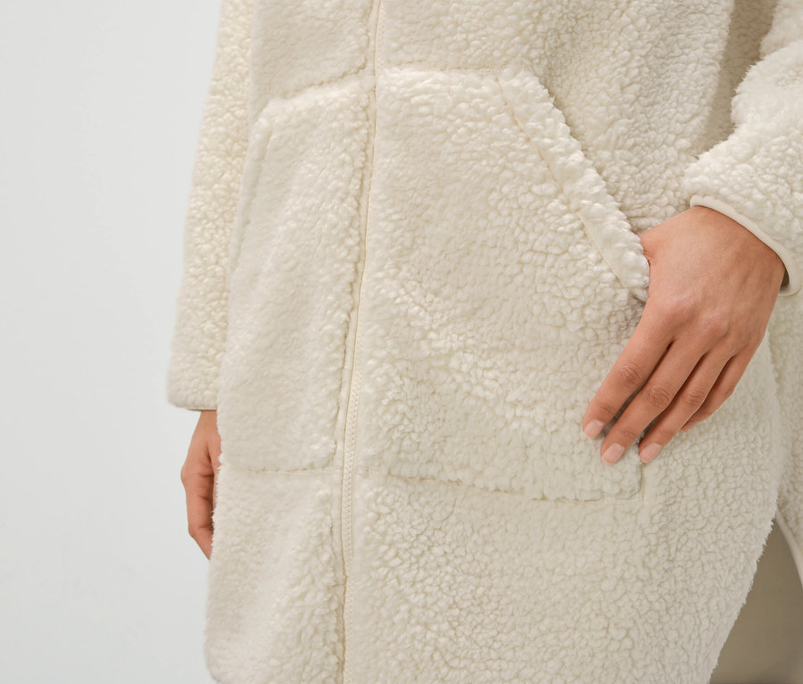 Egy női krémszínű, cipzáras, zsebes műszőrme köntös részlete, kéz a zsebben.