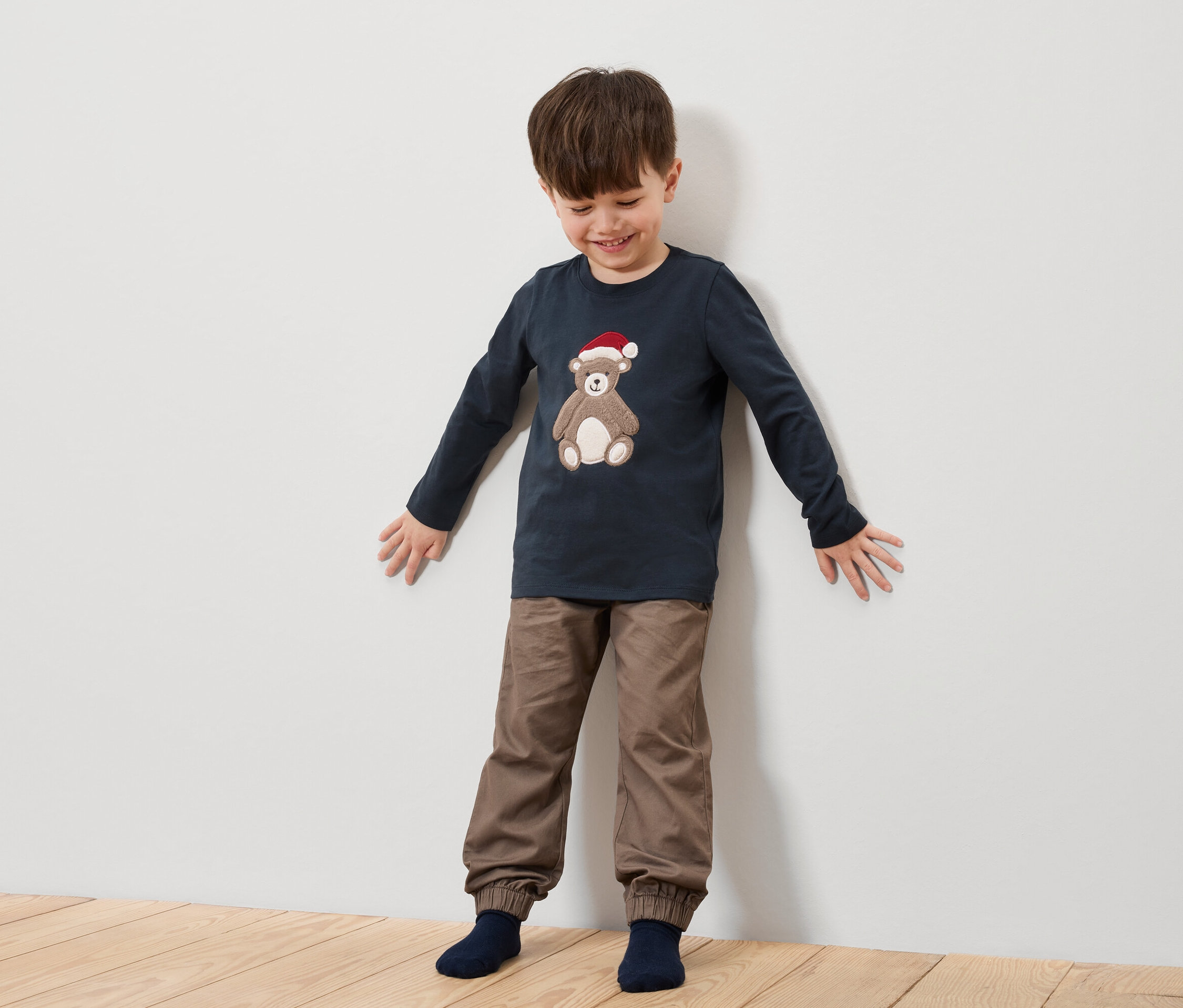 Fiú áll egy fehér fal előtt, sötétkék inget visel, mikulássapkás medvével és barna nadrággal.