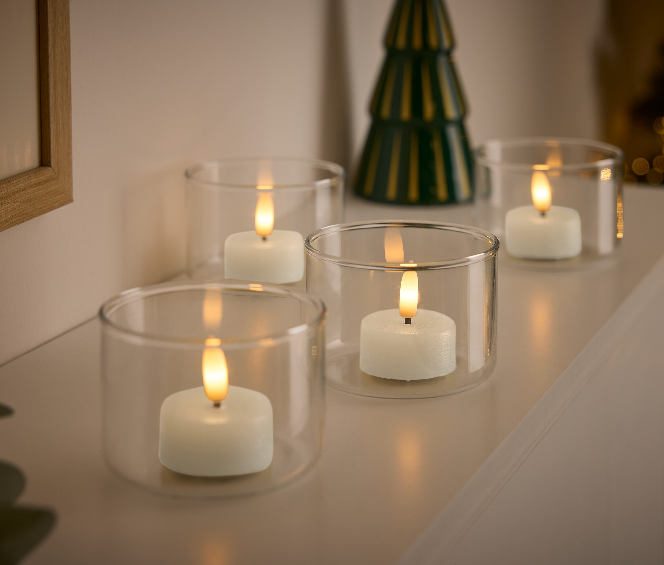 Négy fehér gyertya átlátszó üveg gyertyatartókban, háttérben egy kis zöld karácsonyfával.