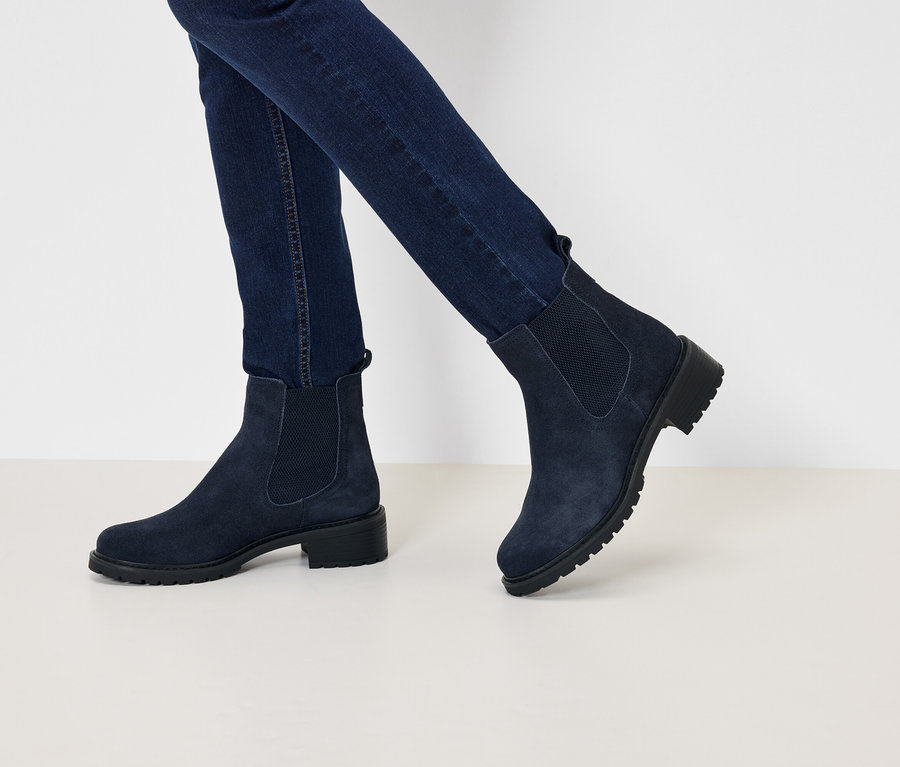 Sötétkék farmerben és tengerészkék velúr, alacsony sarkú Chelsea csizmában lévő lábak sétálnak egy fehér felületen.