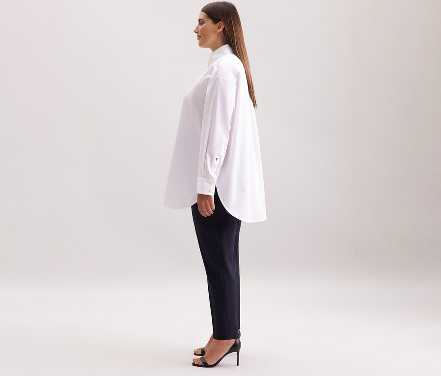Nő visel egy fehér Seidensticker túlméretes hosszú ingblúzt, sötét nadrágot és fekete magas sarkú cipőt.