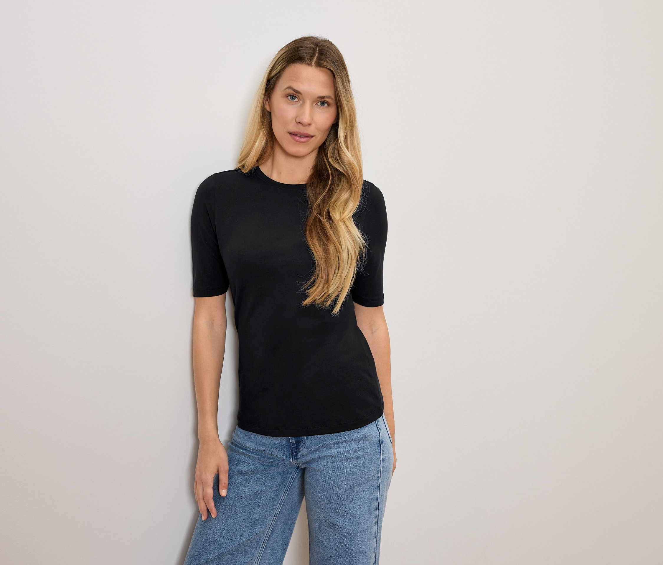 Szőke hajú nő fekete pólóban és kék farmerben pózol.