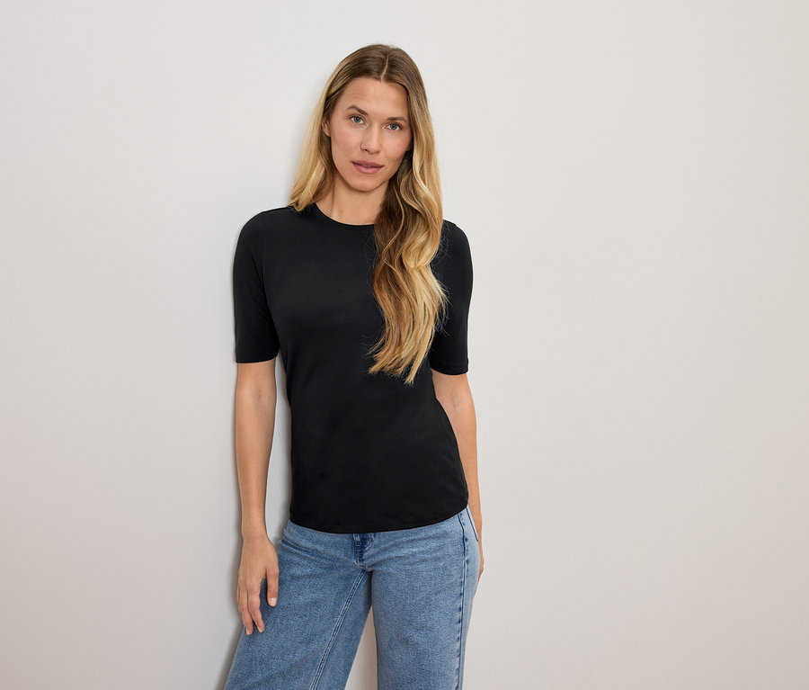 Szőke hajú nő fekete pólóban és kék farmerben pózol.