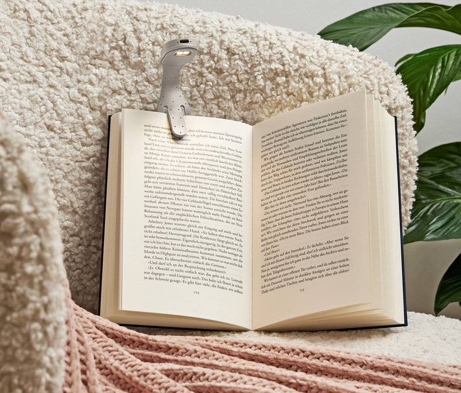 Szürke olvasólámpa van rögzítve egy nyitott könyvre, amely egy puha, krémszínű, rózsaszín díszítésű takarón fekszik.
