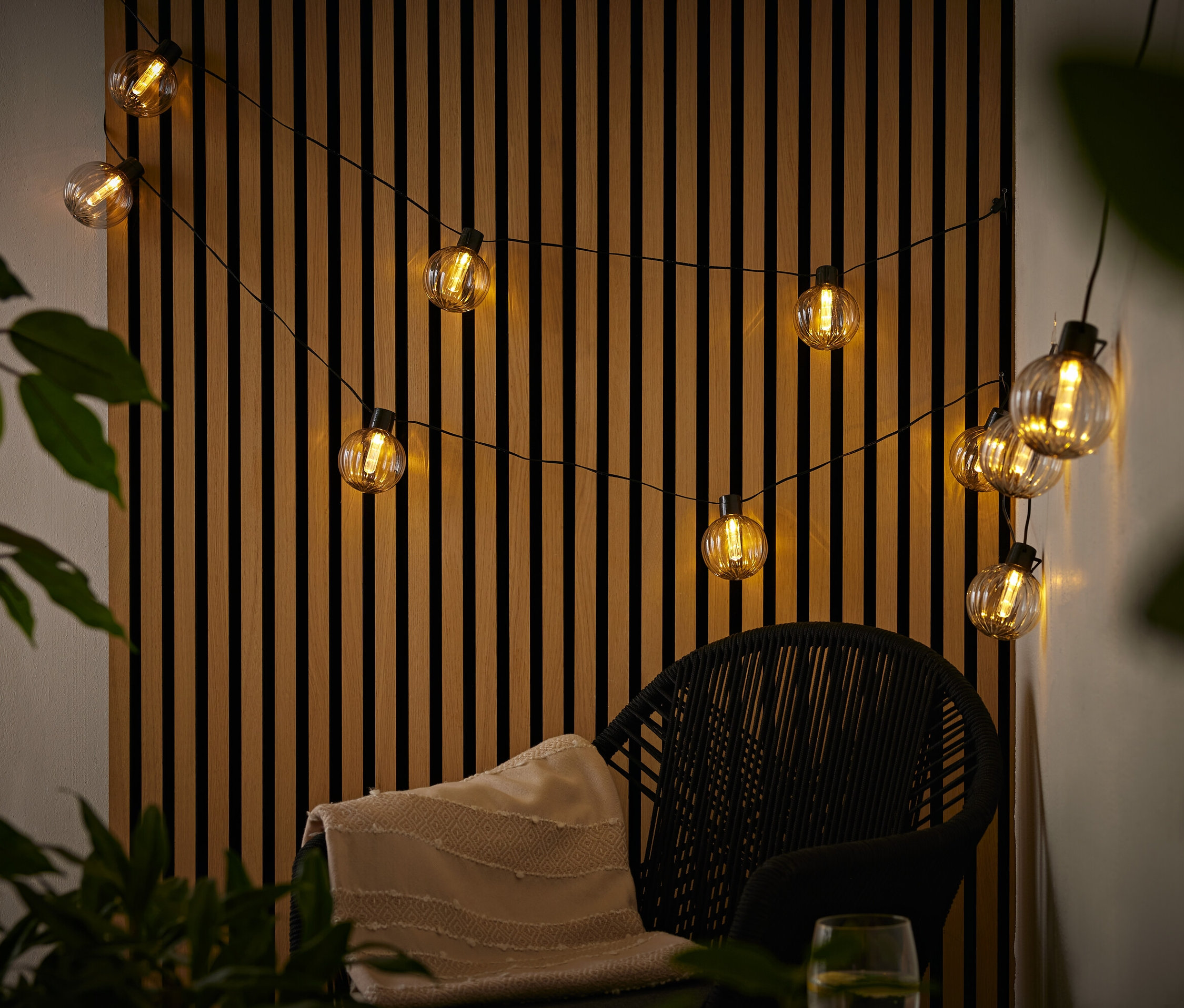 Szolár fényfüzér, hornyos golyók, fekete rattan szék felett, krémszínű takaróval, fa lamellás fal előtt.