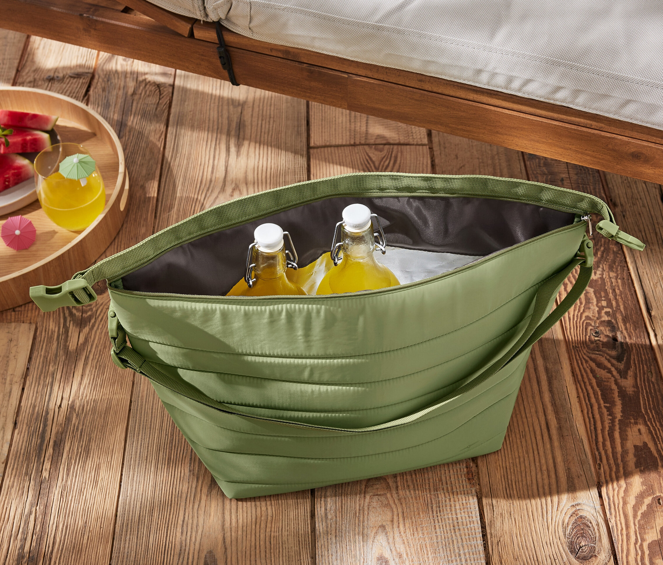 Zöld hűtőtáska palackokkal és görögdinnyével egy faasztalon.