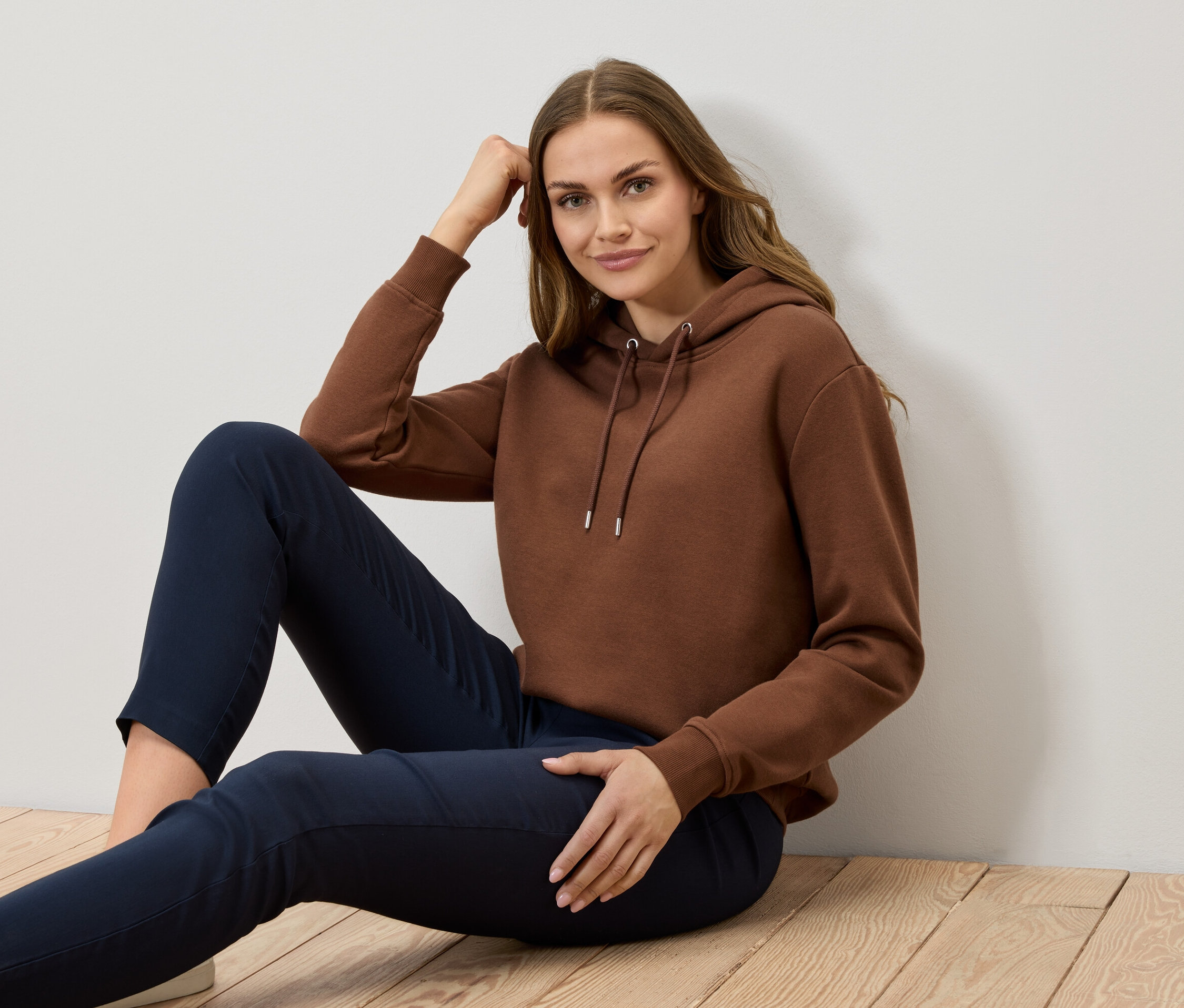 Egy nő ül a fapadlón, barna kapucnis pulóvert és sötétkék nadrágot visel.