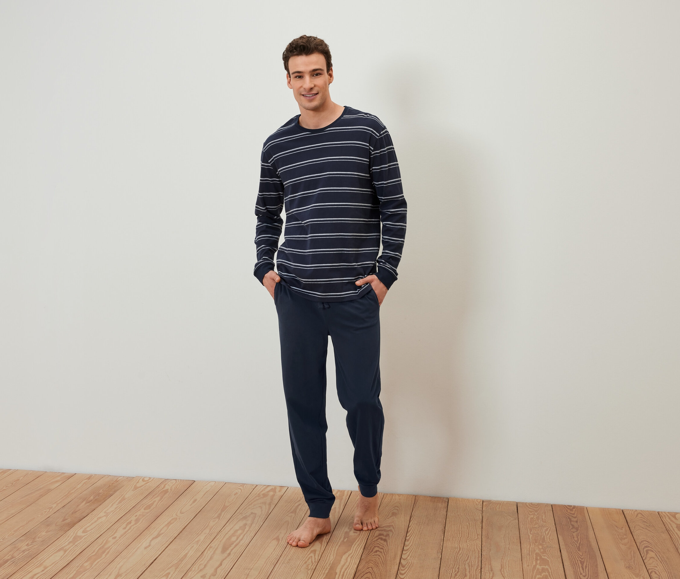 Egy mezítlábas férfi áll, kezével egy sötétkék csíkos pizsama zsebében.