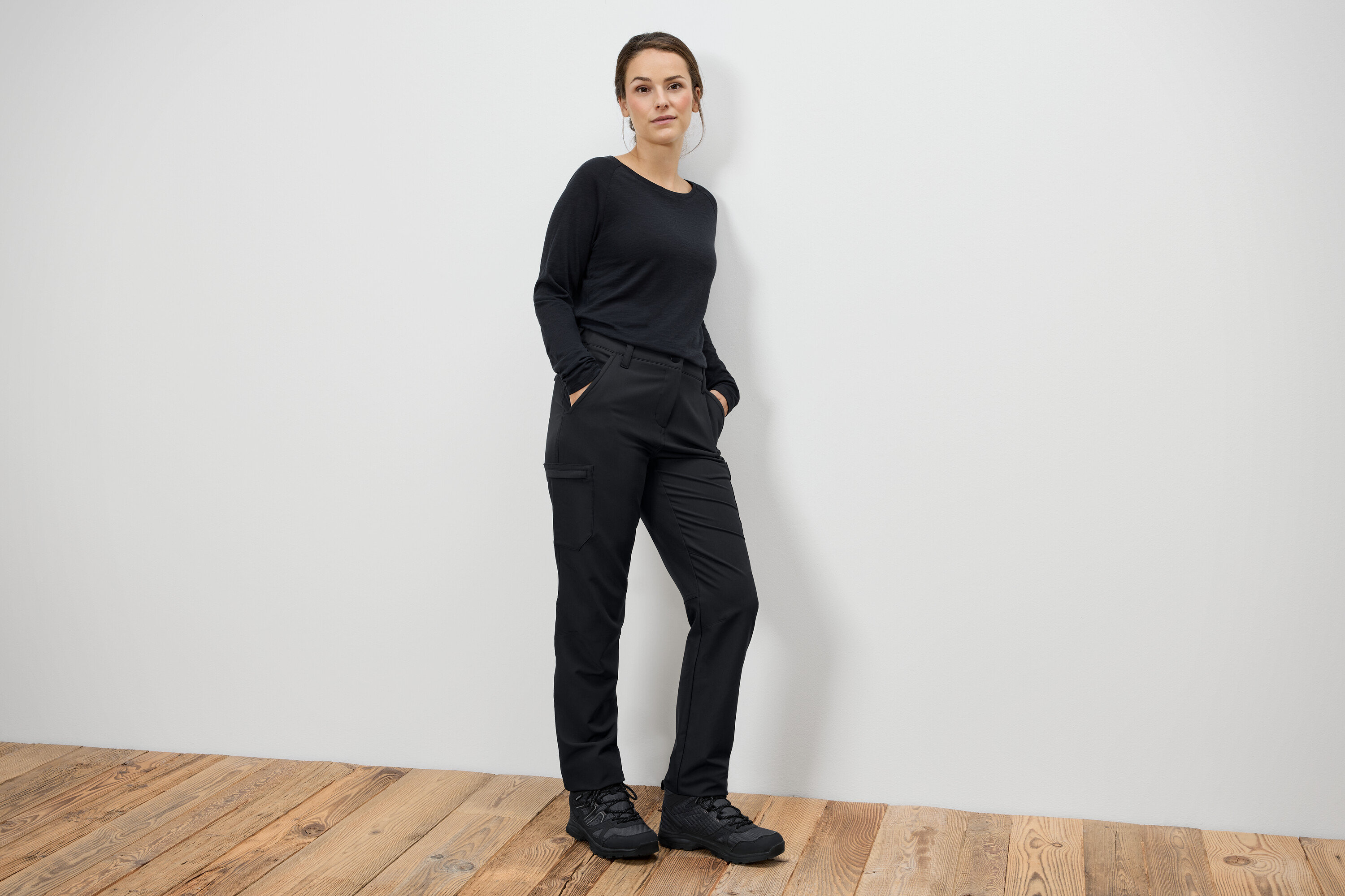 Egy nő áll fekete nadrágban és fekete csizmában, kezét a zsebében tartva.