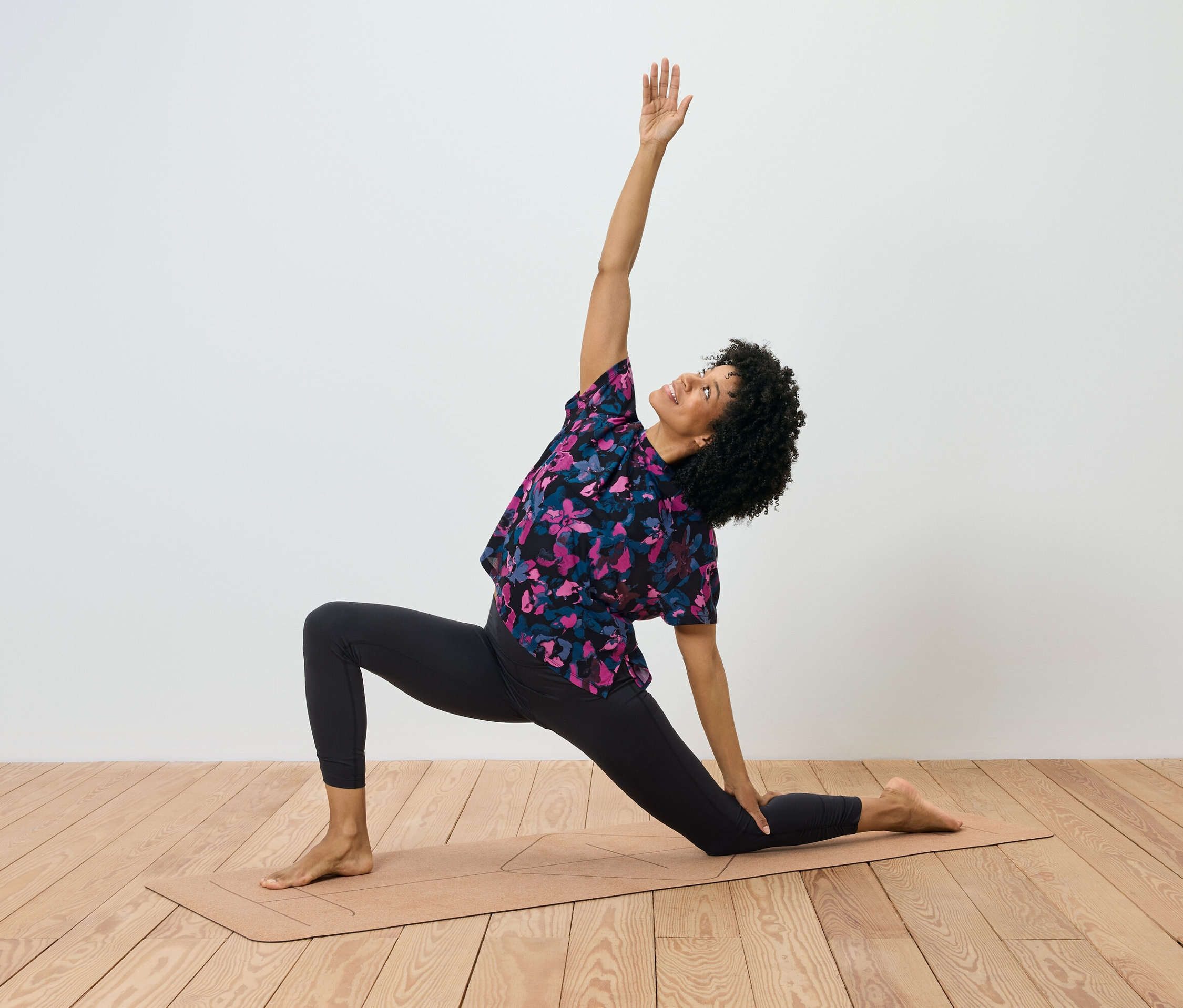 Egy göndör hajú nő jógamatracon gyakorlatozik, a jobb karja felemelve, a bal keze pedig a szőnyeget érinti.