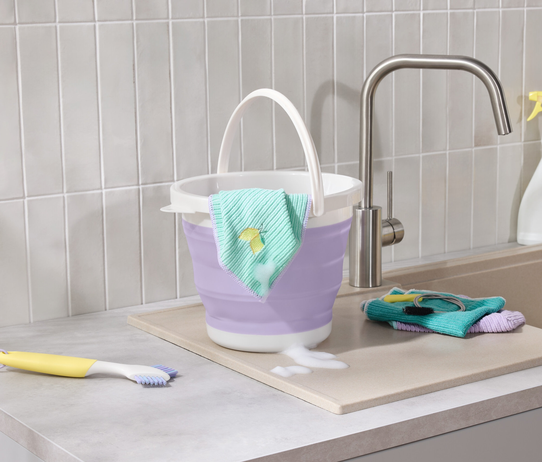 Lila összecsukható vödör citrom hímzésű kendővel a pulton áll a mosogató mellett, ahol egy sárga kefe és rongyok találhatók.