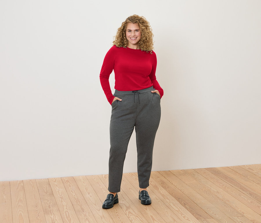 Egy göndör hajú nő áll egy fehér fal előtt, piros hosszú ujjú felsőt és szürke melegítőnadrágot visel.