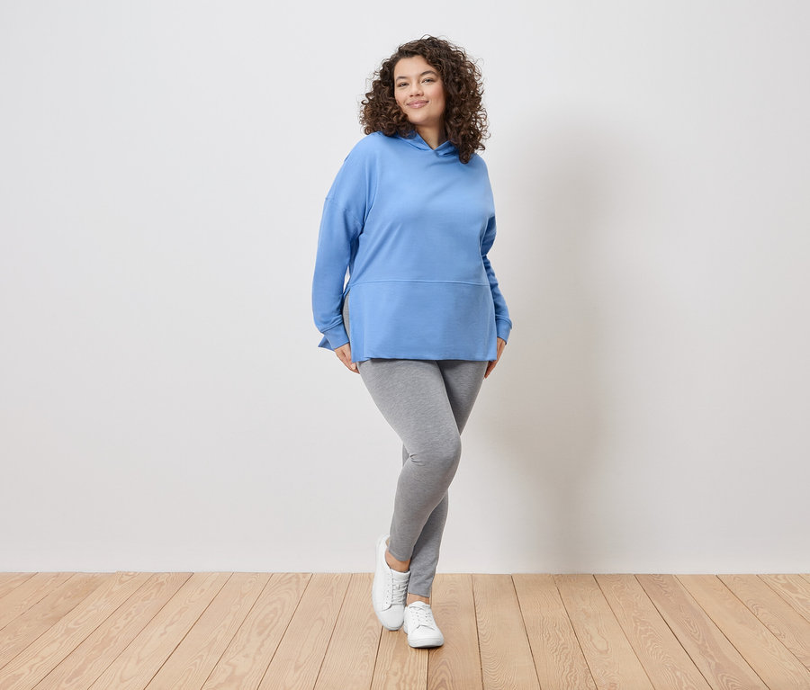 Egy göndör hajú nő áll egy fehér fal előtt, kék kapucnis pulóvert, szürke leggingset és fehér tornacipőt visel.